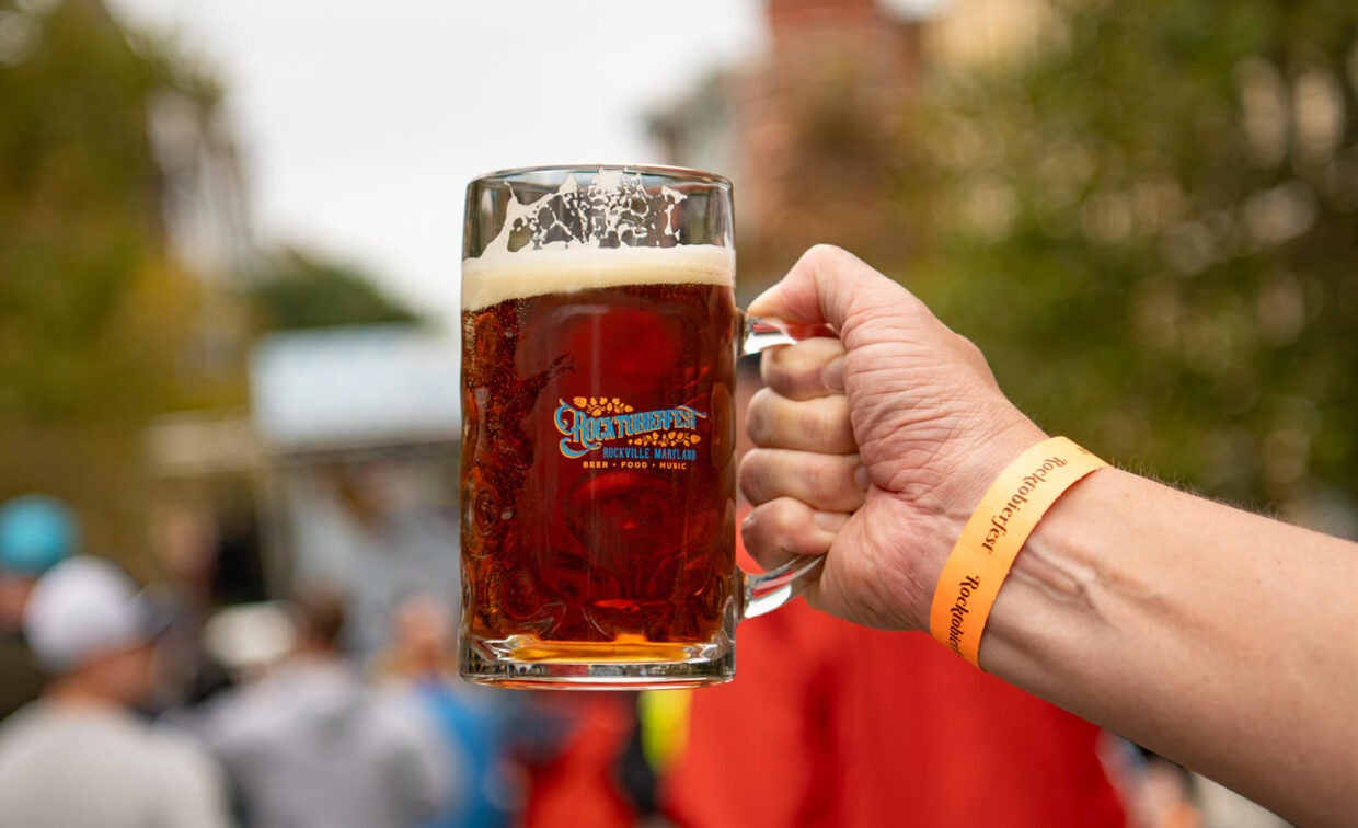 Hand holding a glass mug of beer with the Rocktobierfest logo and a matching event wristband.
