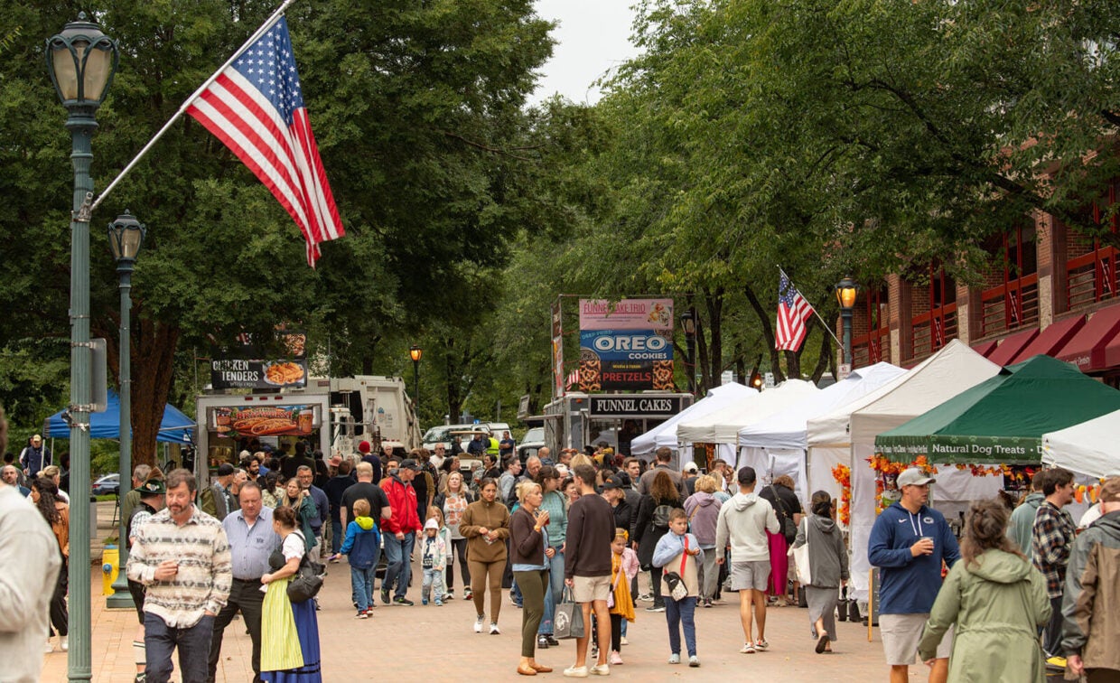 A street festival with food vendor trucks, white tents, and people walking along a tree-lined brick path under American flags.