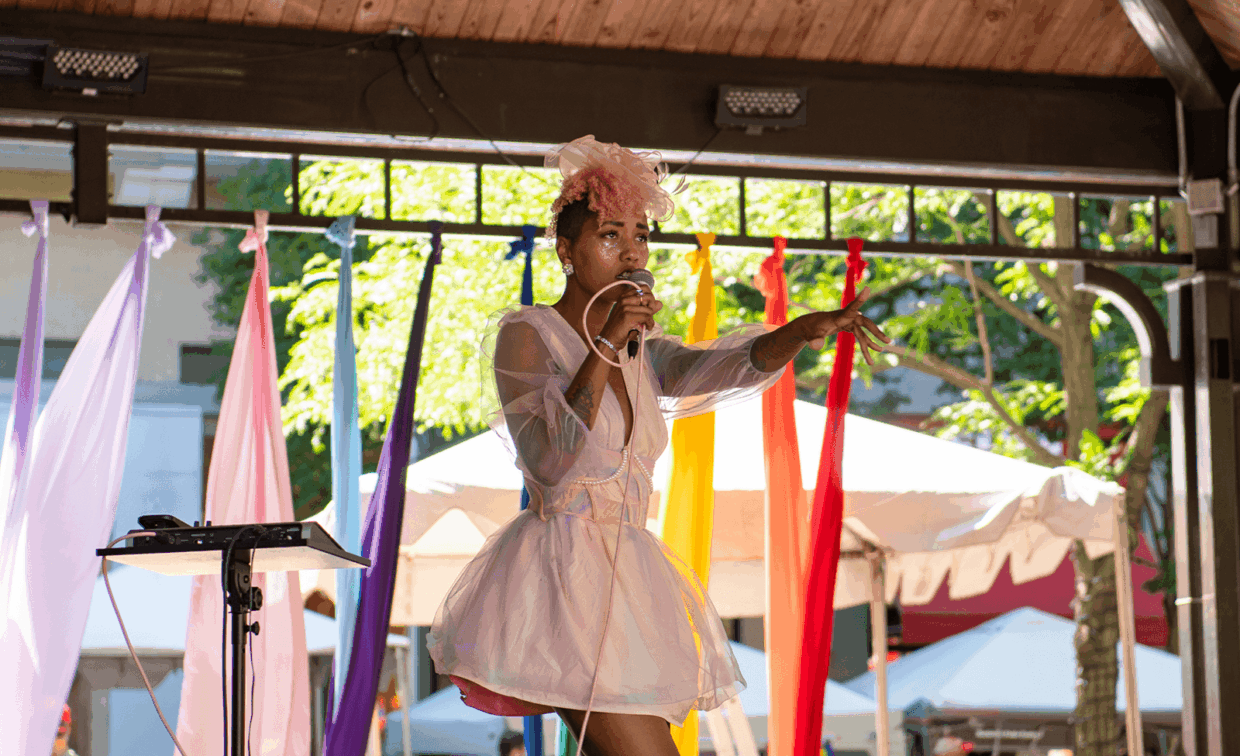 A performer in a white dress and headpiece sings into a microphone on an outdoor stage decorated with colorful fabric streamers.