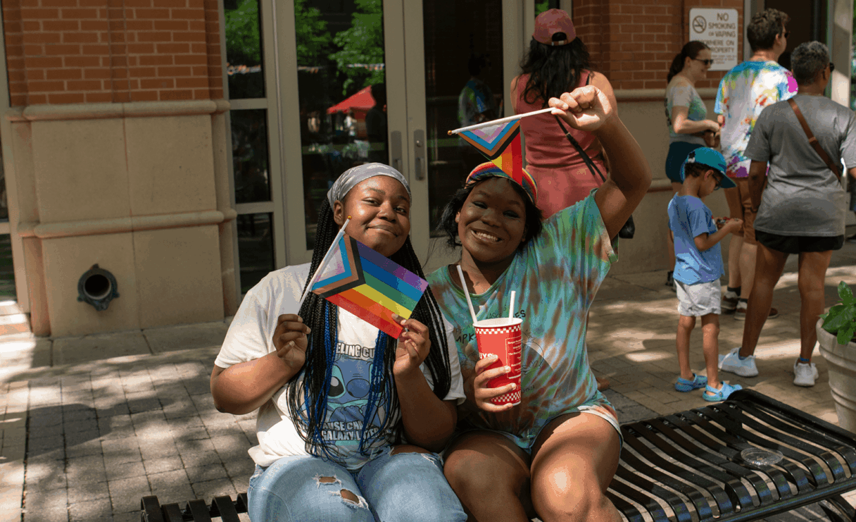Two people sit on a bench outdoors, holding small Progress Pride flags.