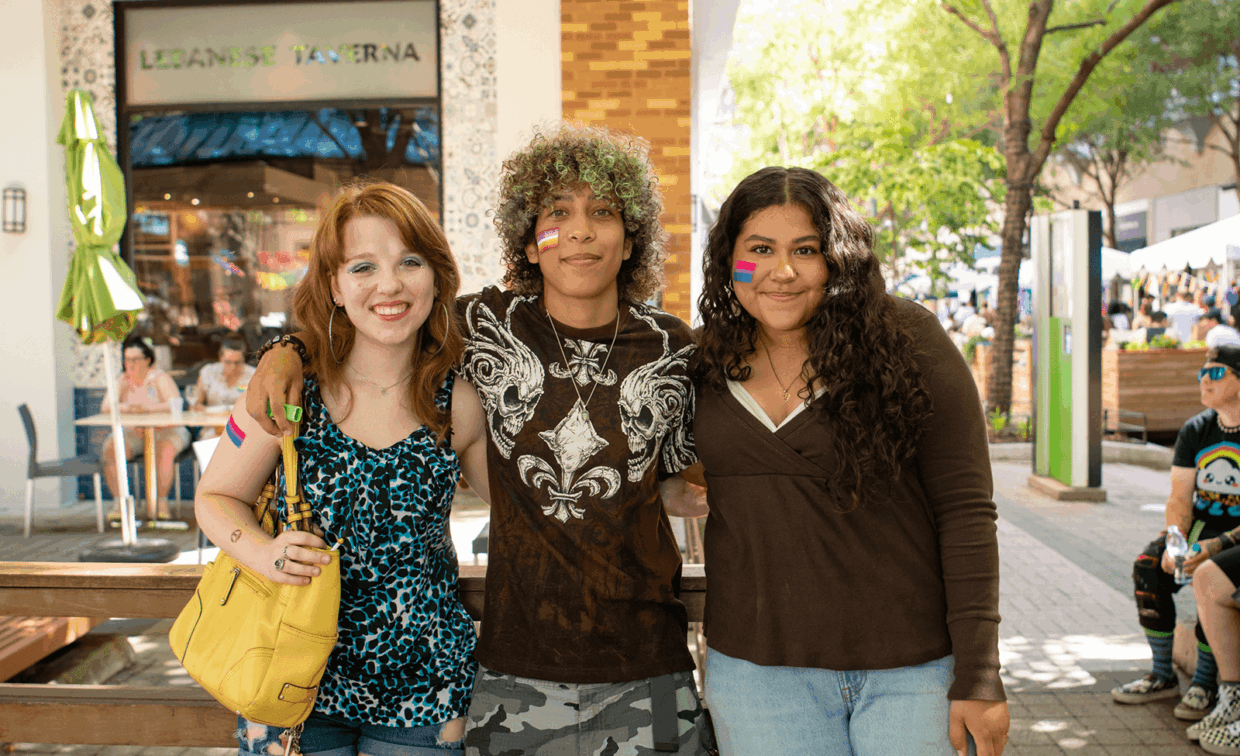 Three people stand together outdoors, each wearing a small bisexual pride flag sticker on their cheek.