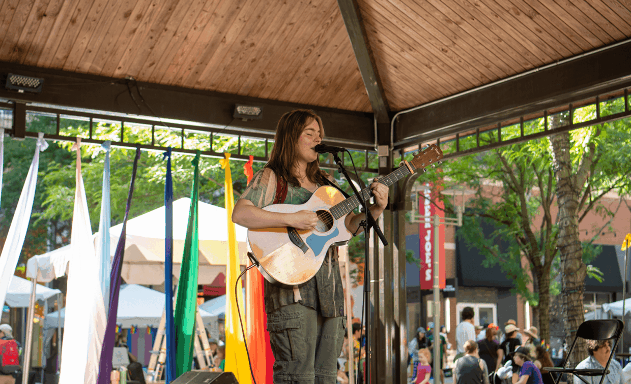 A musician playing an acoustic guitar and singing into a microphone on a stage decorated with colorful fabric streamers.