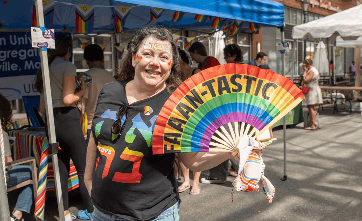 A smiling woman with face paint holds a rainbow fan with the text “FAAAN-TASTIC” at an outdoor event.