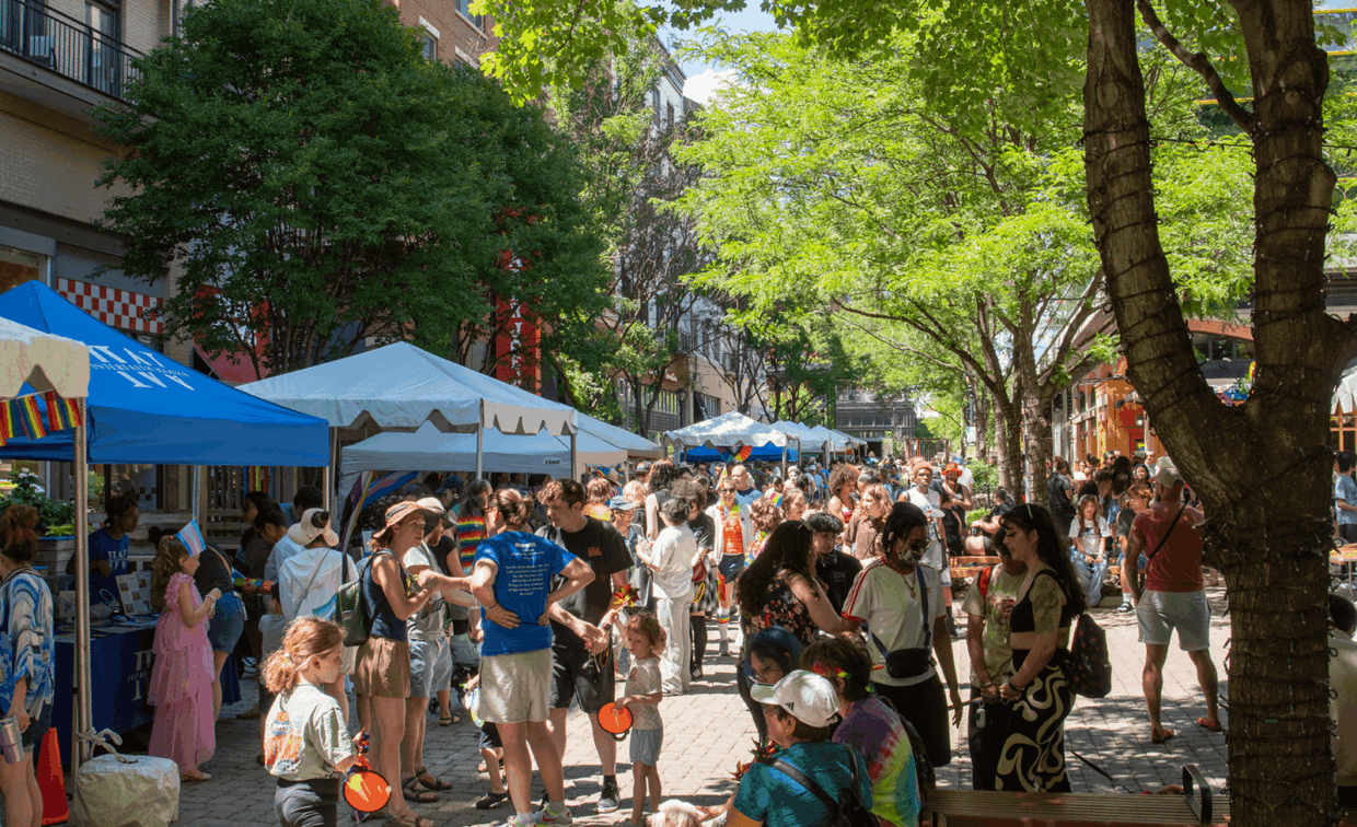 A crowded outdoor street festival with vendor tents lining a brick pathway shaded by trees.