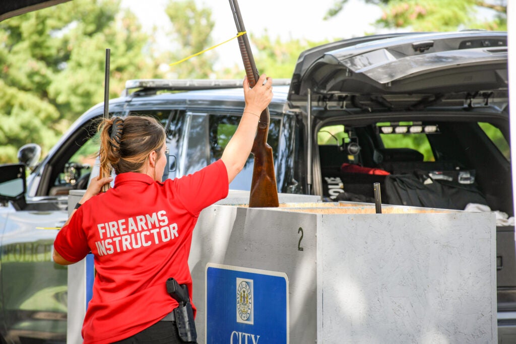 A firearms instructor in a red polo shirt placing a shotgun into a wooden crate at a shooting range.